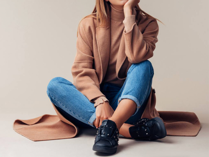 woman sitting down with jeans, camel colored turtleneck, camel coat, and black sneakers