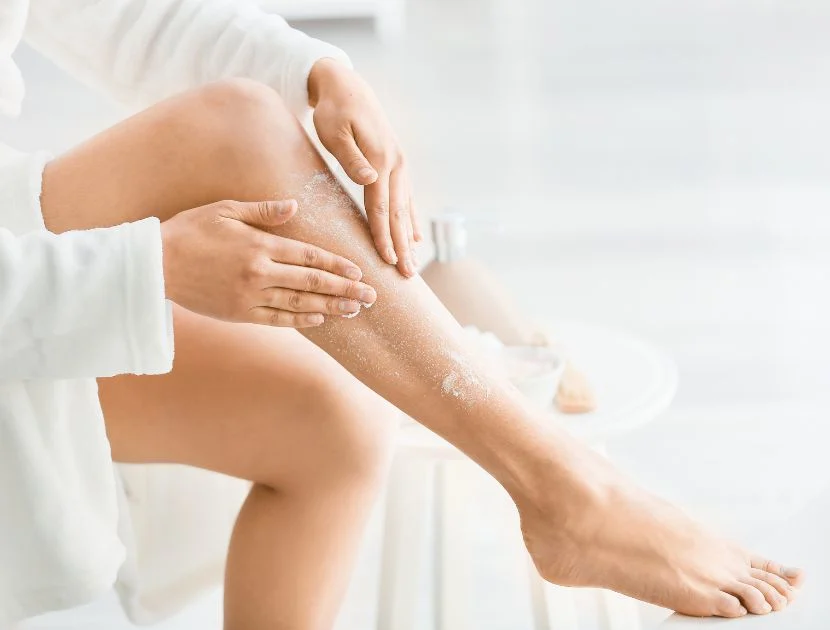 Woman exfoliating her legs with body scrub while wearing a white bathrobe, demonstrating a full body skincare routine for smoother skin after 40